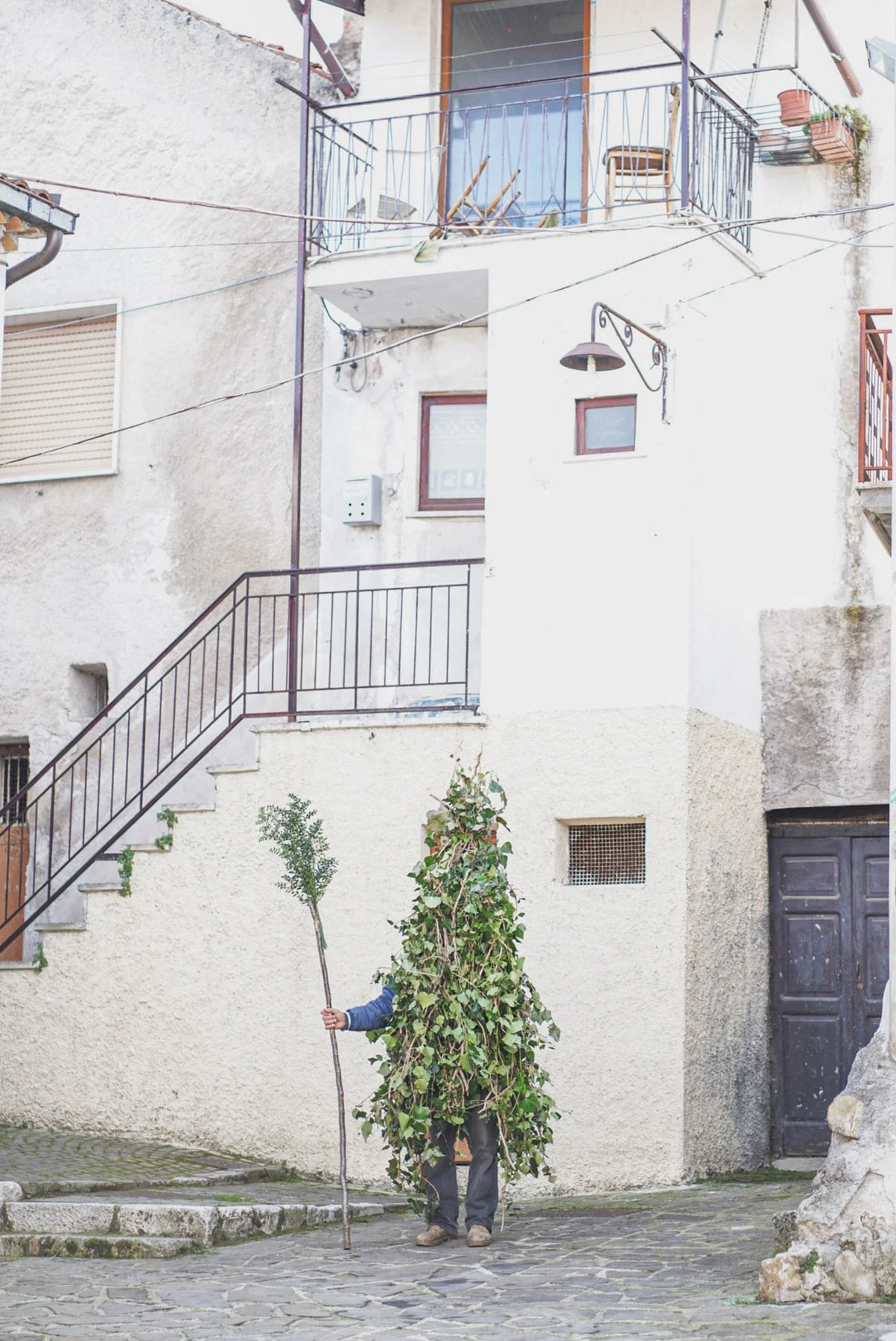 A photograph of a man wearing a “Rumita” outfit (made from tree branches) while standing under the stairs of a white home in Satriano village, Italy.